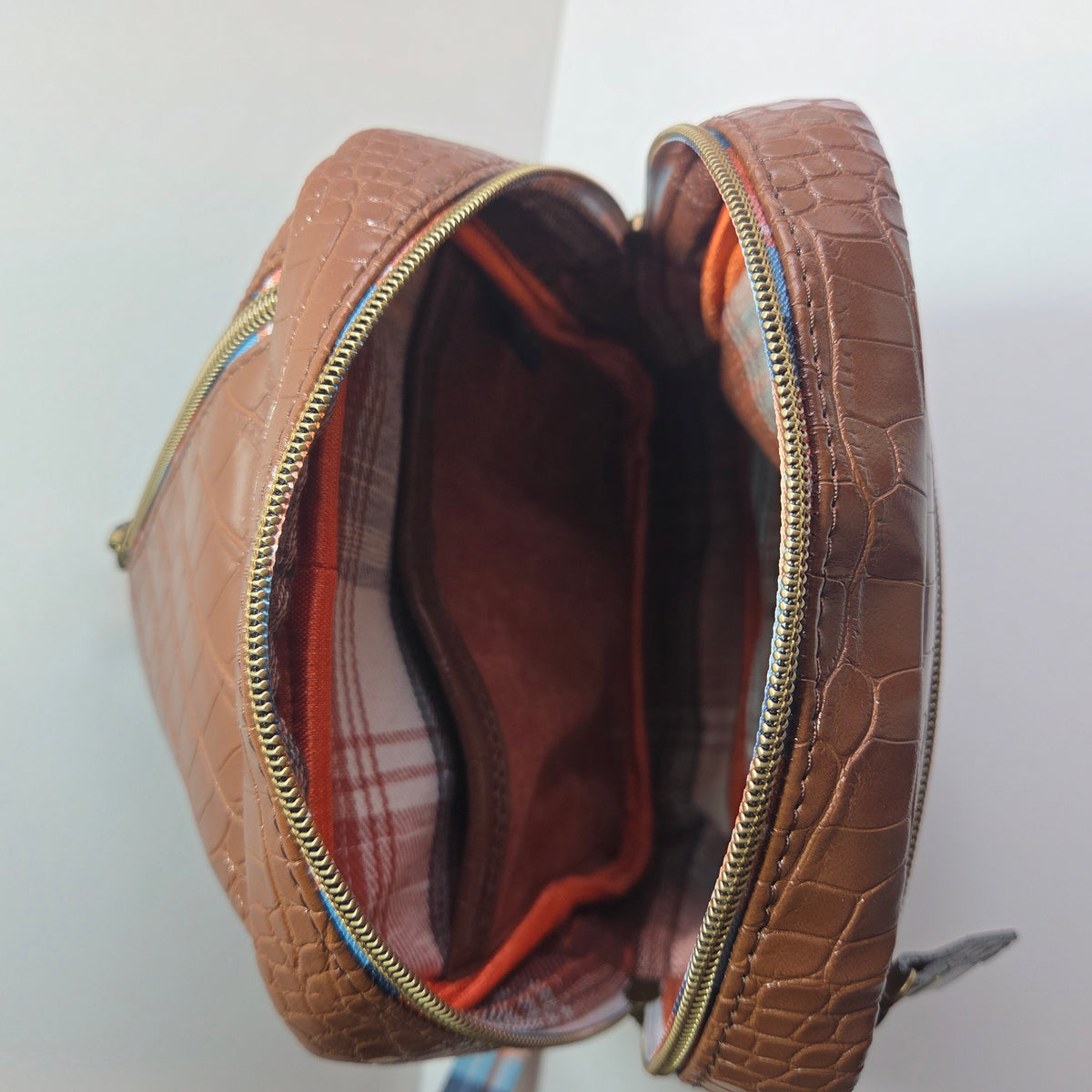 A brown crocodile-embossed zippered Sling bag shown open, revealing a brown plaid lining, slip pocket and orange binding.