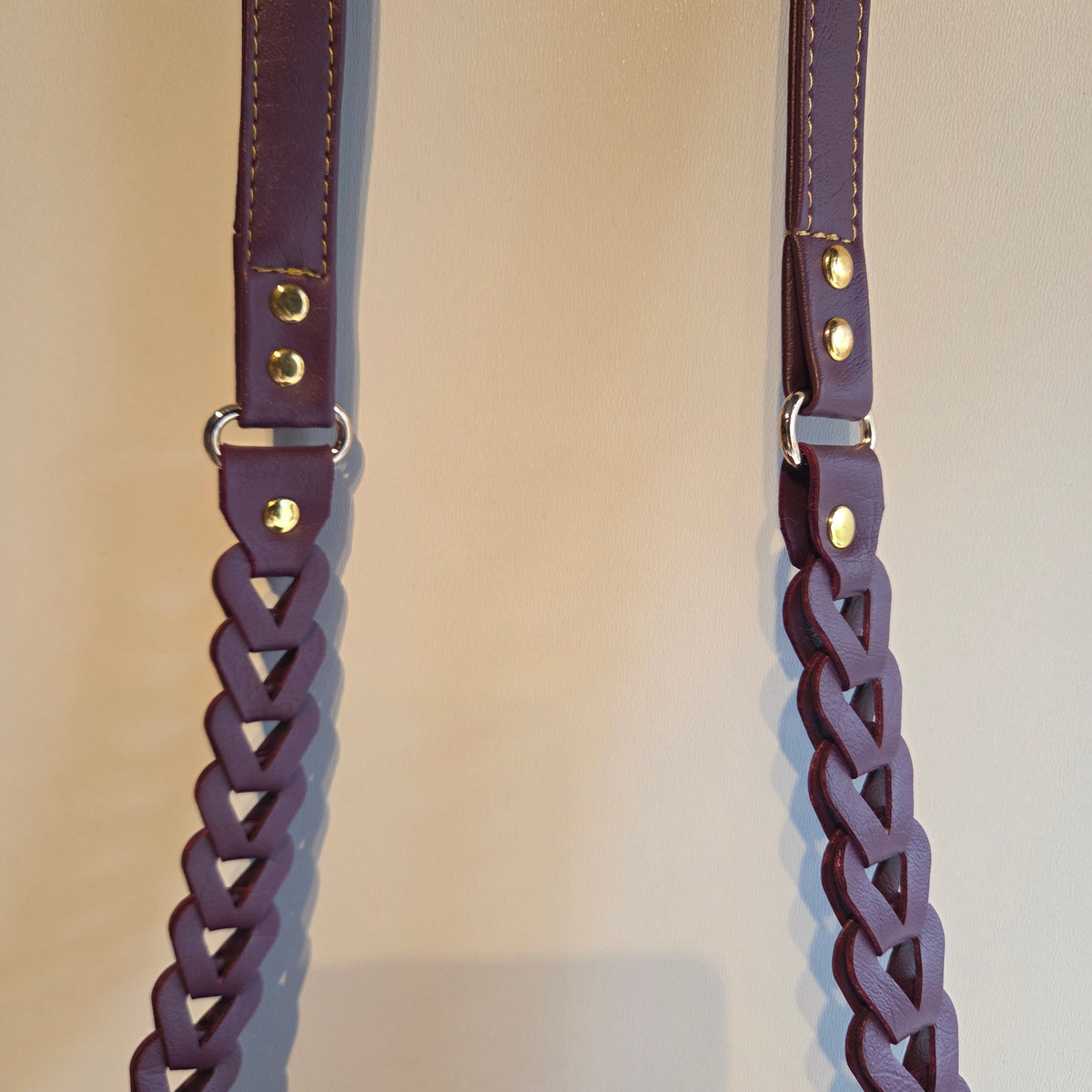 A close-up of a dark purple braided faux leather strap with gold-tone metal rivets and gold stitching, shown against a plain beige background.