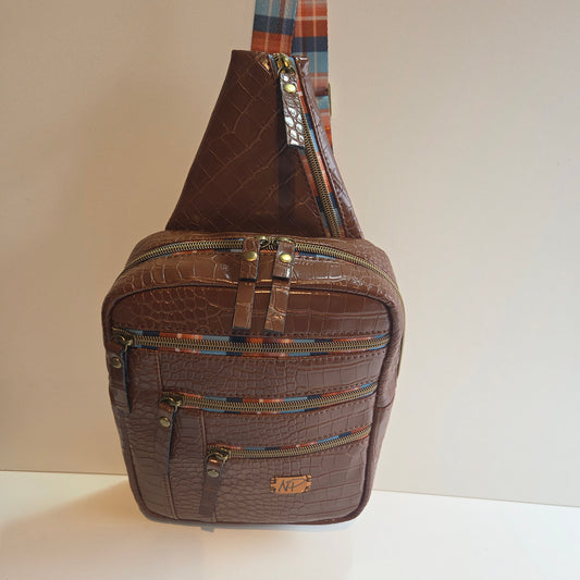 A brown crocodile-embossed leather sling bag with multiple zippered compartments and a colourful striped patterned strap.