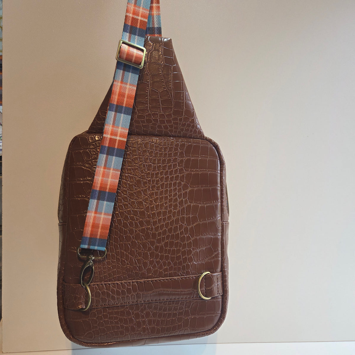 A brown crocodile-embossed faux leather sling bag with an, adjustable plaid strap in orange, blue, and white, shown against a plain light background.