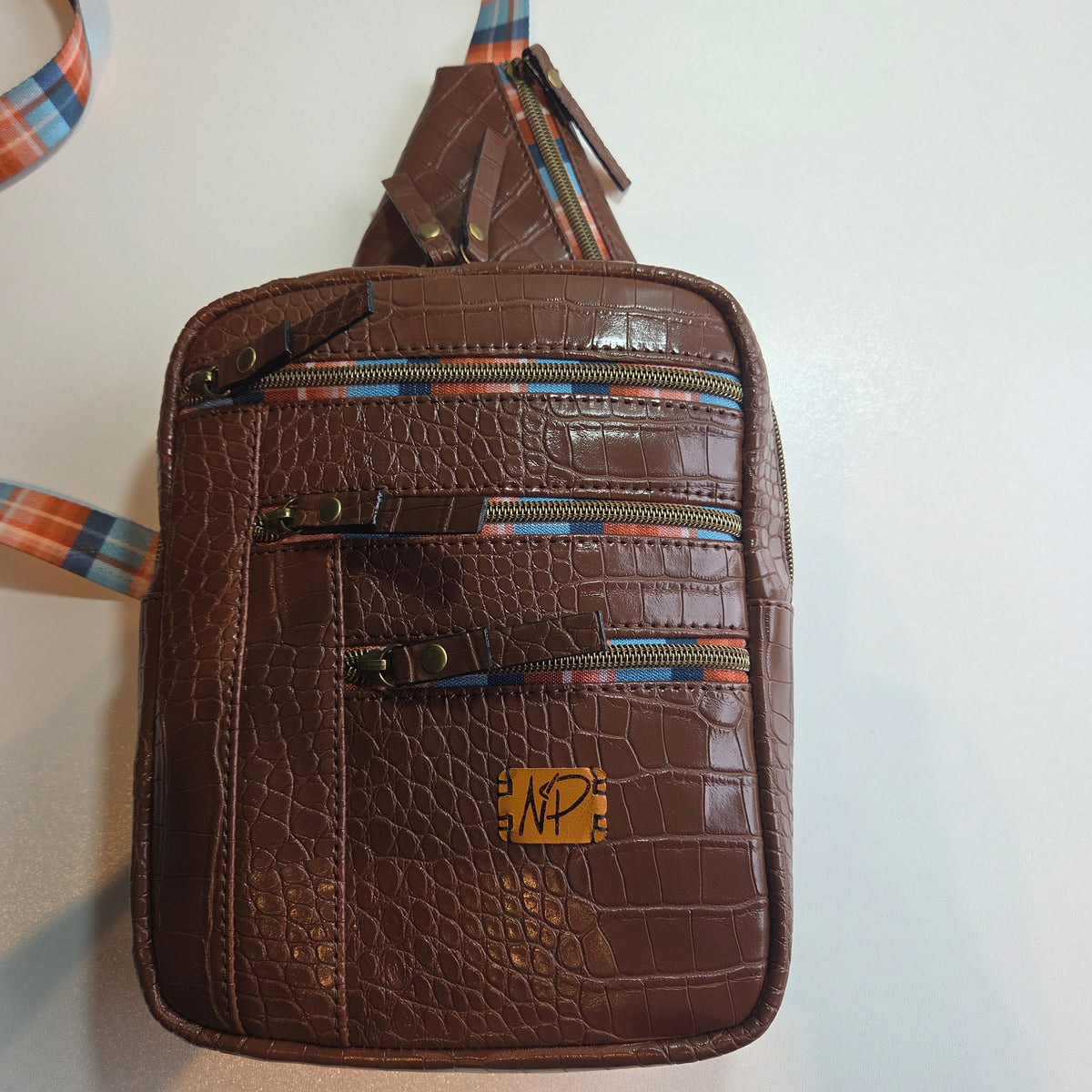 A brown crocodile-embossed sling bag with multiple zippered compartments and a plaid patterned strap, shown on a plain light background.