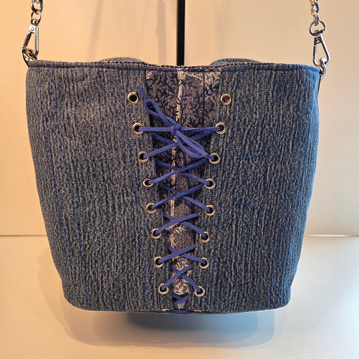 A denim bag with a lace-up back detail in blue cord and silver-tone metal eyelets, shown against a plain light background.