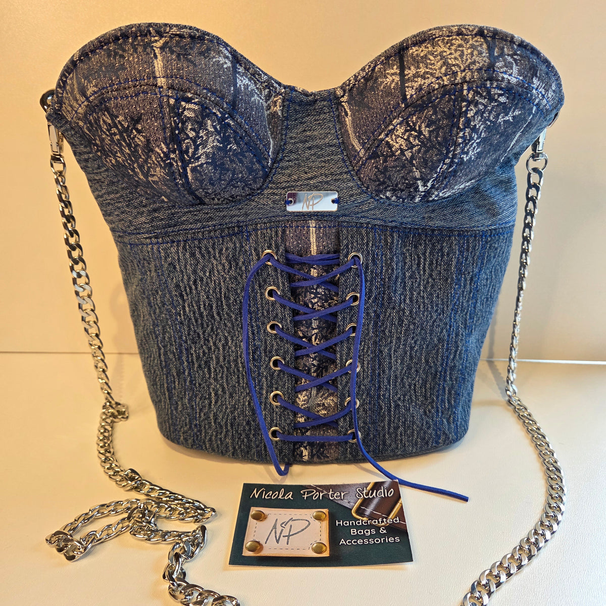 A blue denim corset-style handbag with lace-up front, silver chain strap, and a small brand card in front on a light beige background.