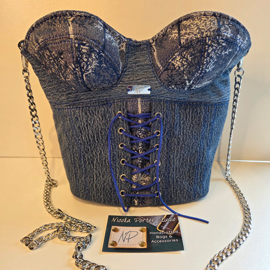 A blue denim corset-style handbag with lace-up front, silver chain strap, and a small brand card in front on a light beige background.