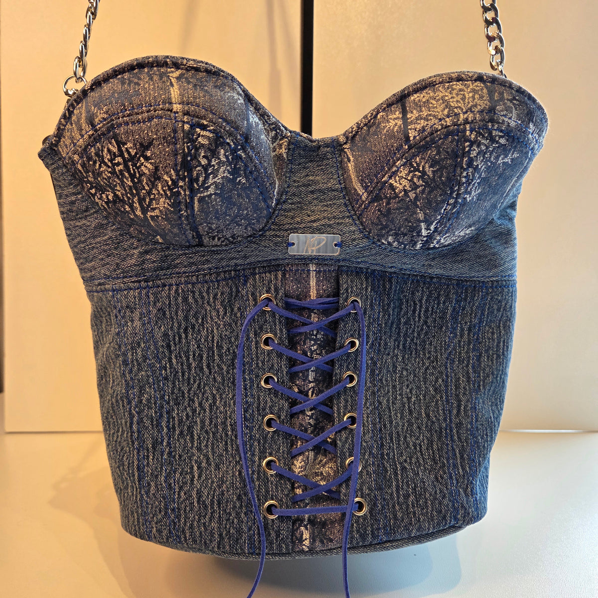 A corset-style shoulder bag made from distressed denim, featuring a lace-up front, silver-tone chain straps, and a small mirrored plaque on the front.
