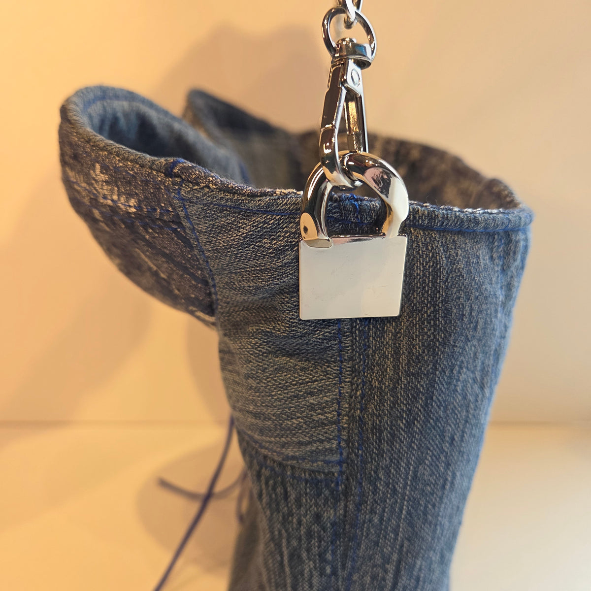 A close-up of a blue denim bag with a silver metal strap connector and chain strap attached, set against a plain beige background.