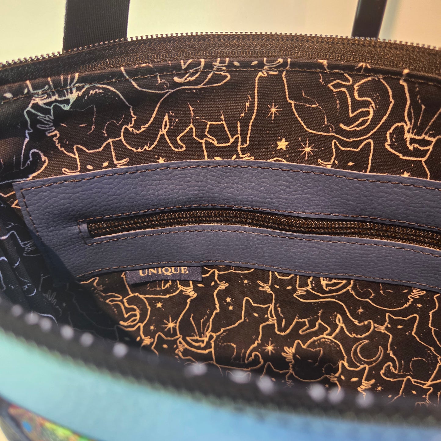 Lining of a bag showing a blue zip pocket overlay and label. Fabric is black with white cat outlines and stars