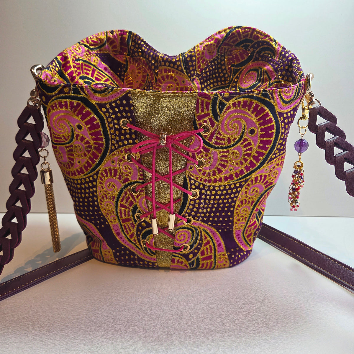 A handmade corset-style bucket bag with a purple, pink, and gold paisley patterned fabric body, pink laces, and a purple brown braided shoulder strap.