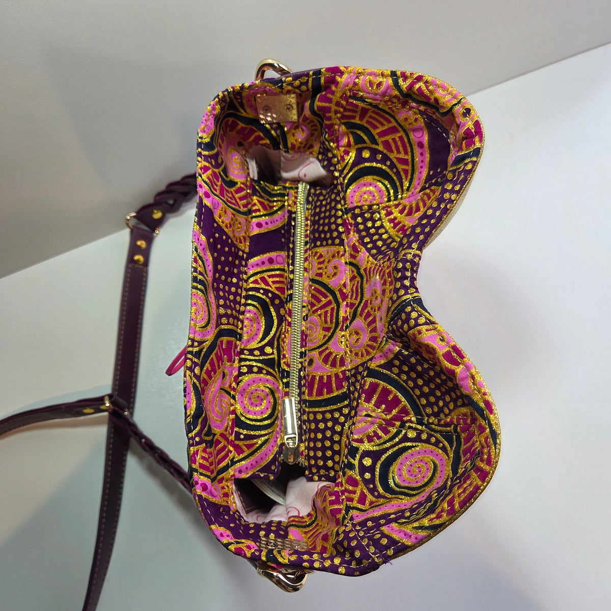 A close-up of a handbag's closed top showing a vibrant, paisley and floral pattern in purple, pink and gold. A gold zipper acts as the closure, and a dark purple shoulder strap with gold studs is visible in the foreground against a white background.