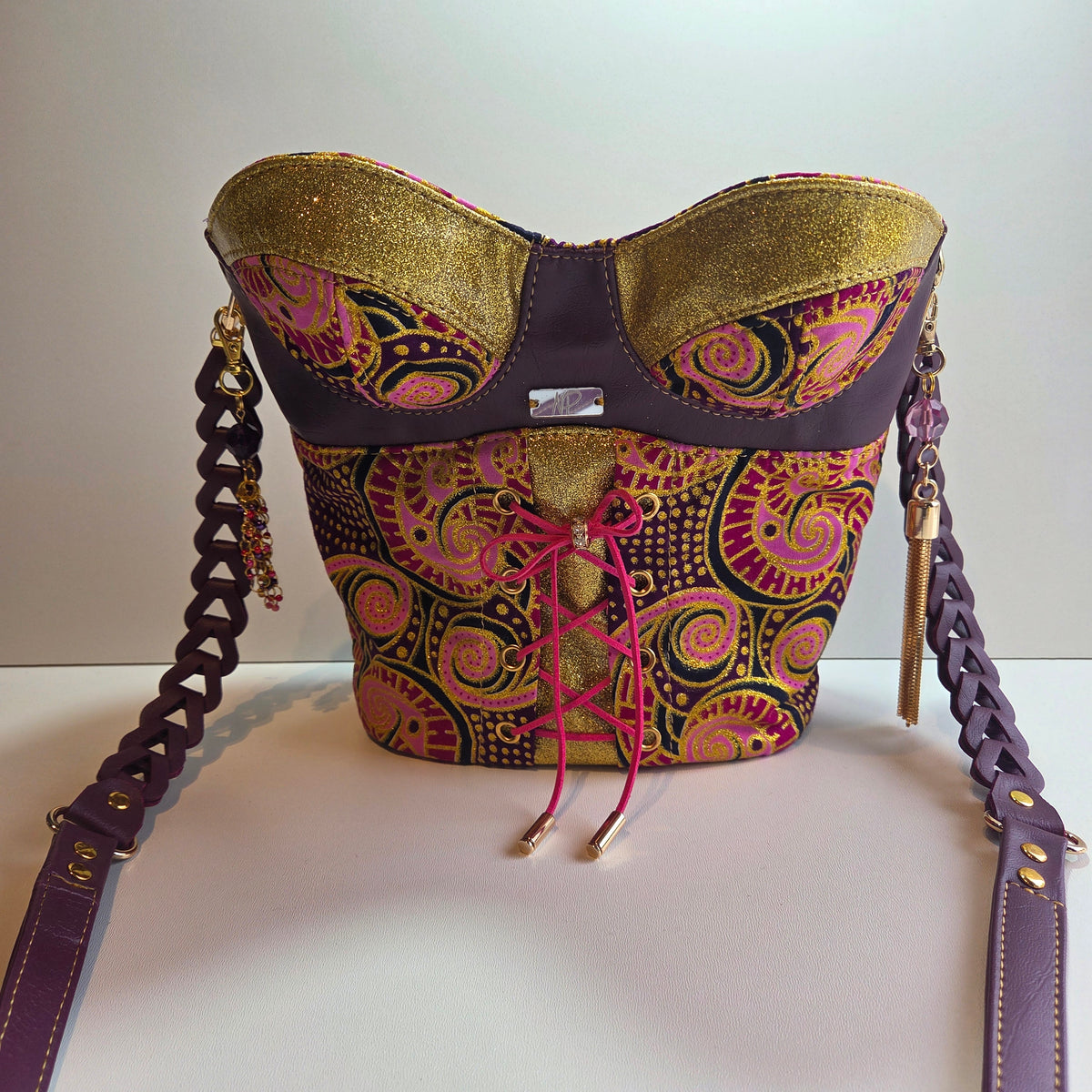 A handbag with a sculptural bust-like silhouette, featuring a purple braided shoulder strap, gold glitter bust panels, and a central panel with a pink, purple, and black paisley pattern. The bag is shown against a plain off-white background.
