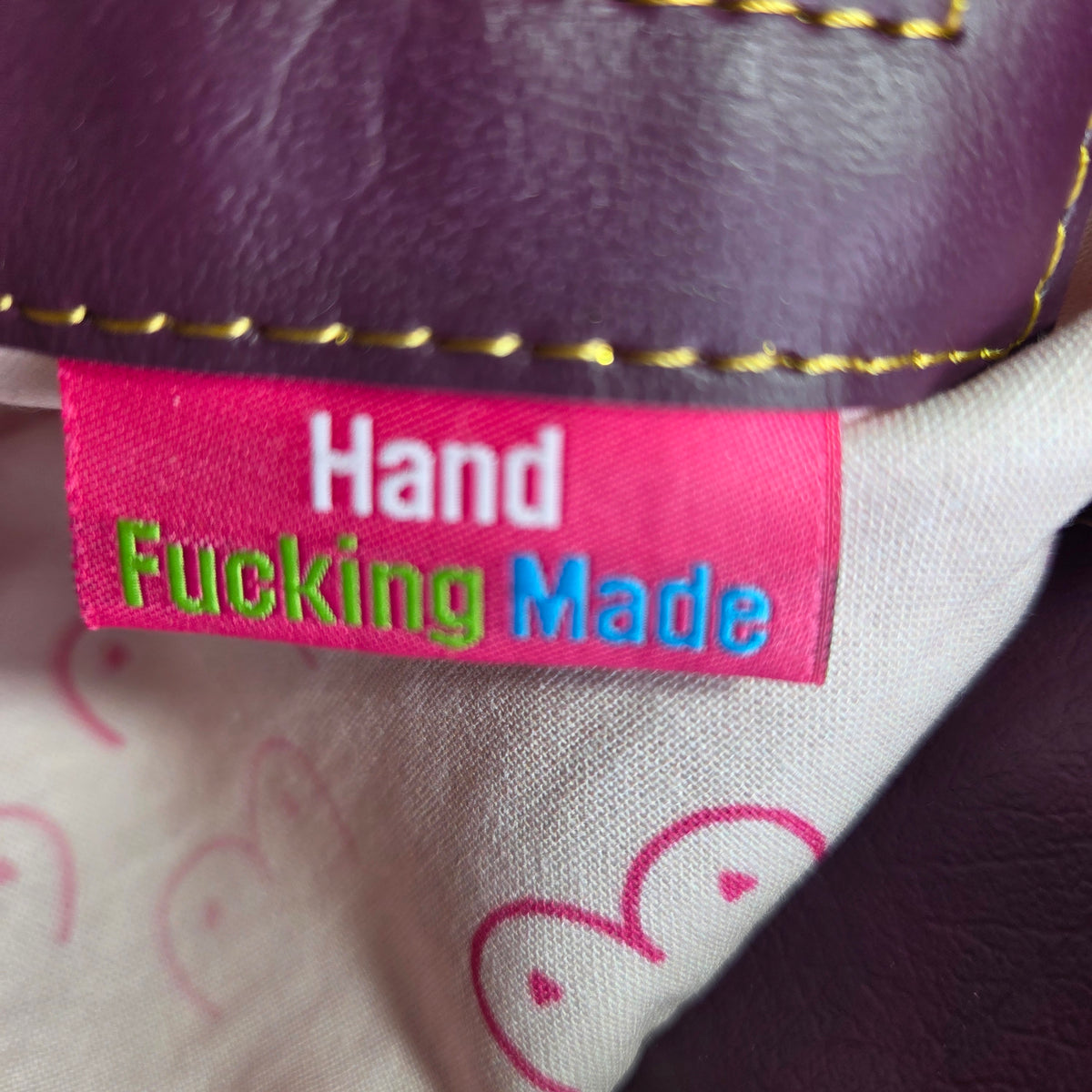 Close-up of a pink fabric tag reading 'Hand Fucking Made' in white, blue, and green embroidery, on the inside of a handmade bag with gold stitching and a boob patterned fabric lining.