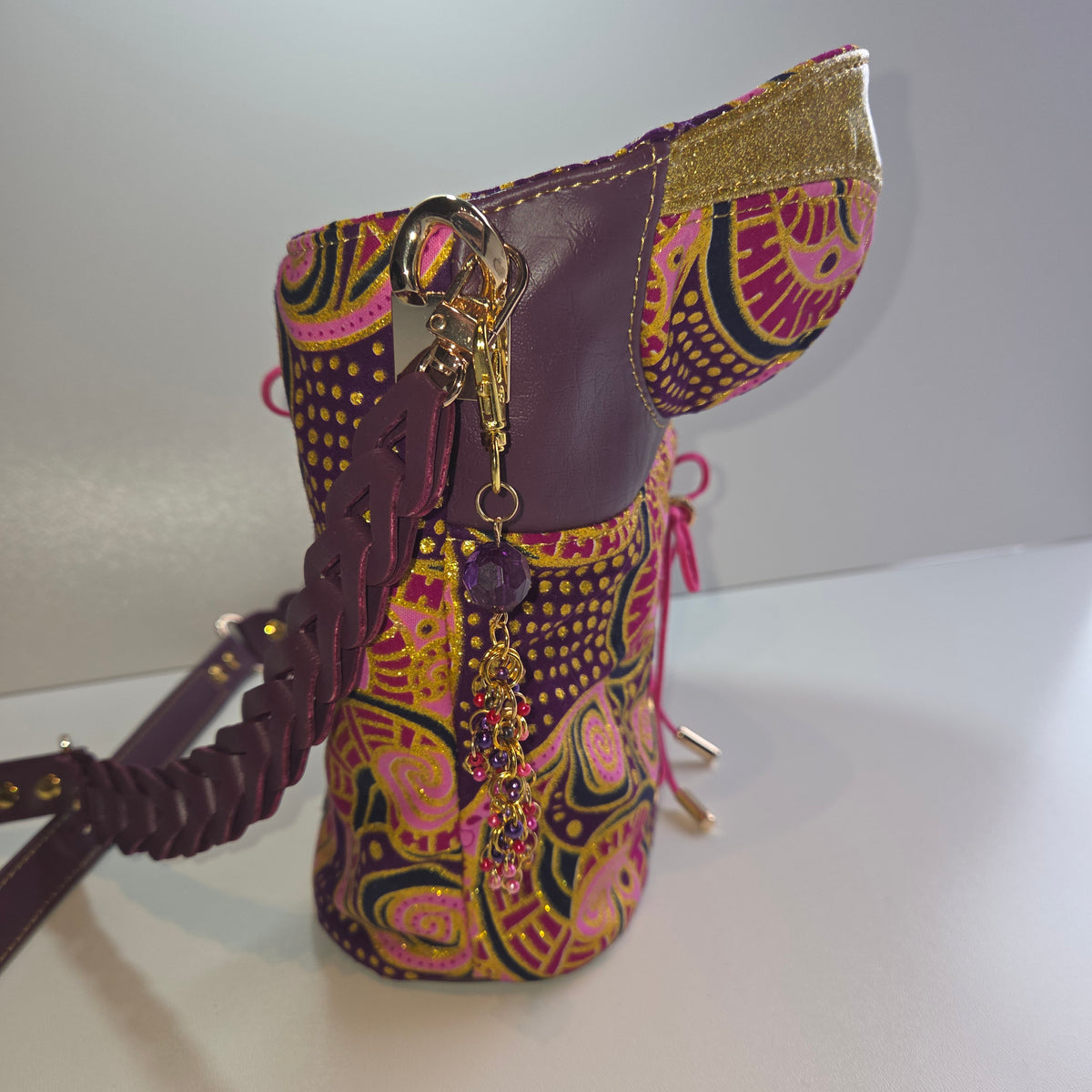 A side view of basque shaped handbag with a purple strap and trim, decorated with a colourful paisley pattern in pink, yellow, and green, and accented with gold-tone hardware.