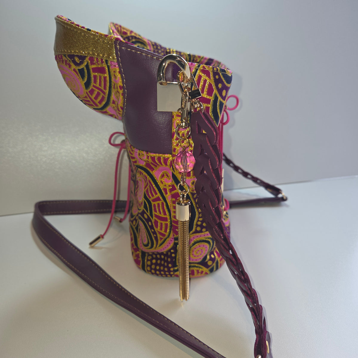A side view of basque shaped handbag with a purple strap and trim, decorated with a colourful paisley pattern in pink, yellow, and green, and accented with gold-tone hardware.