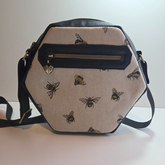 A handmade hexagonal shoulder bag with a cream background printed with black and yellow bees, featuring black faux-leather trim and a vertical zippered pocket on the back, with heart shaped zip pull.