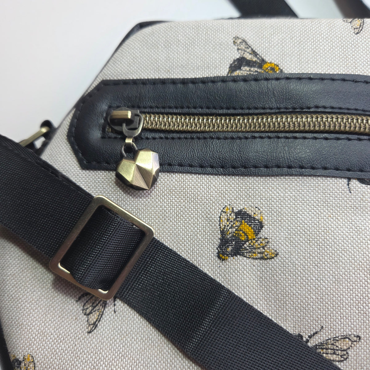 Close-up of a cream canvas bag panel with a black faux-leather zipper panel, brass finish zipper, bee print, and a black adjustable strap with a metal buckle.