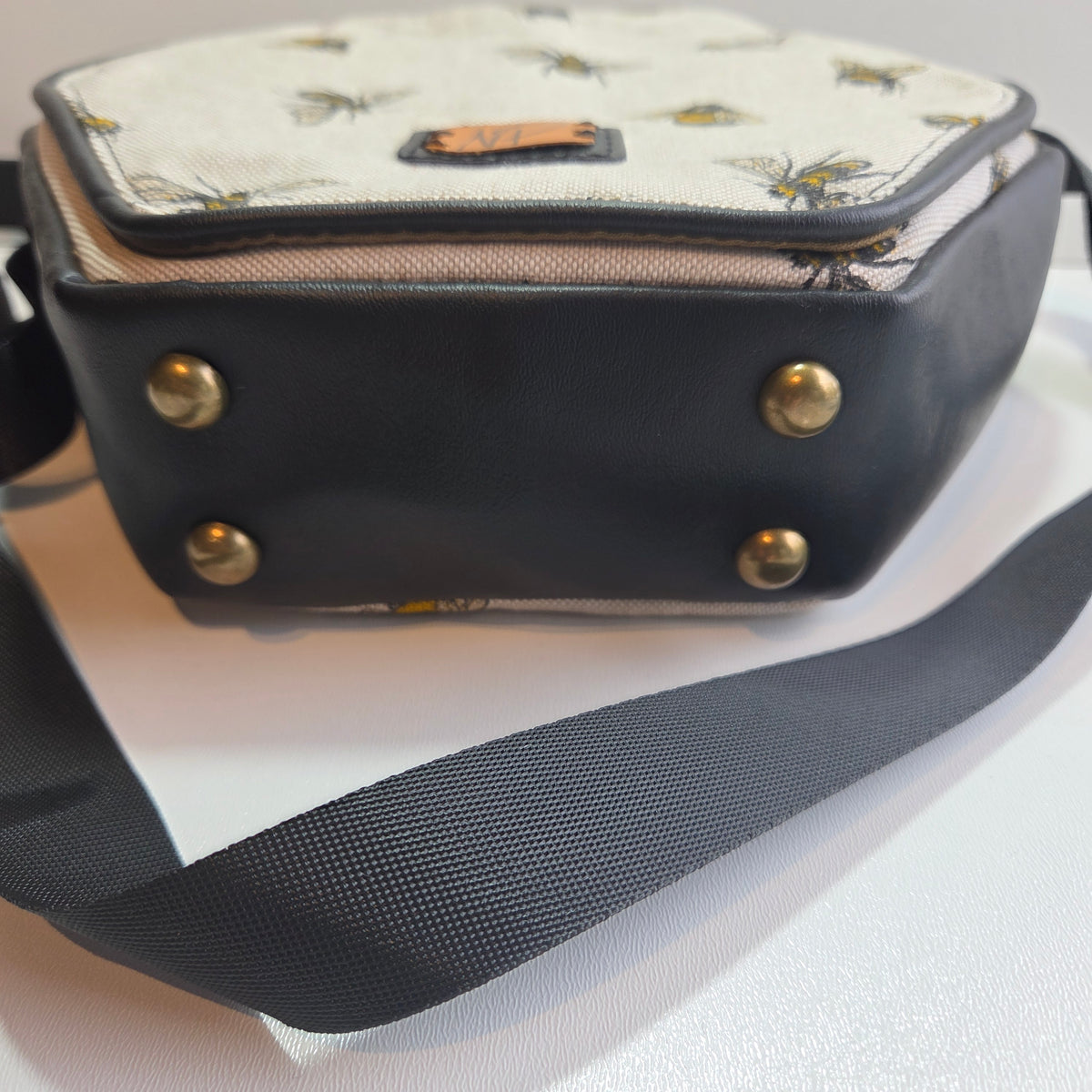 A close-up of a small bag base with a black vinyl body, brass-tone feet studs, and a cream canvas panel printed with yellow and black bees. A black webbing strap is visible against a plain white background.