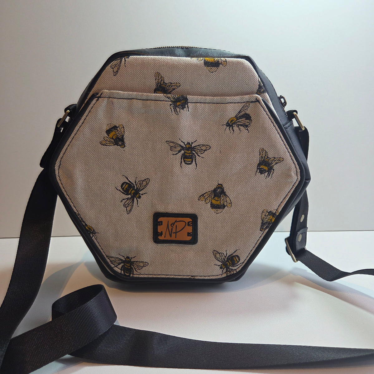 A small handmade hexagonal shoulder bag with a cream background printed with black and yellow bee motifs, featuring black trim, a black strap, and a small brown logo patch on the front.