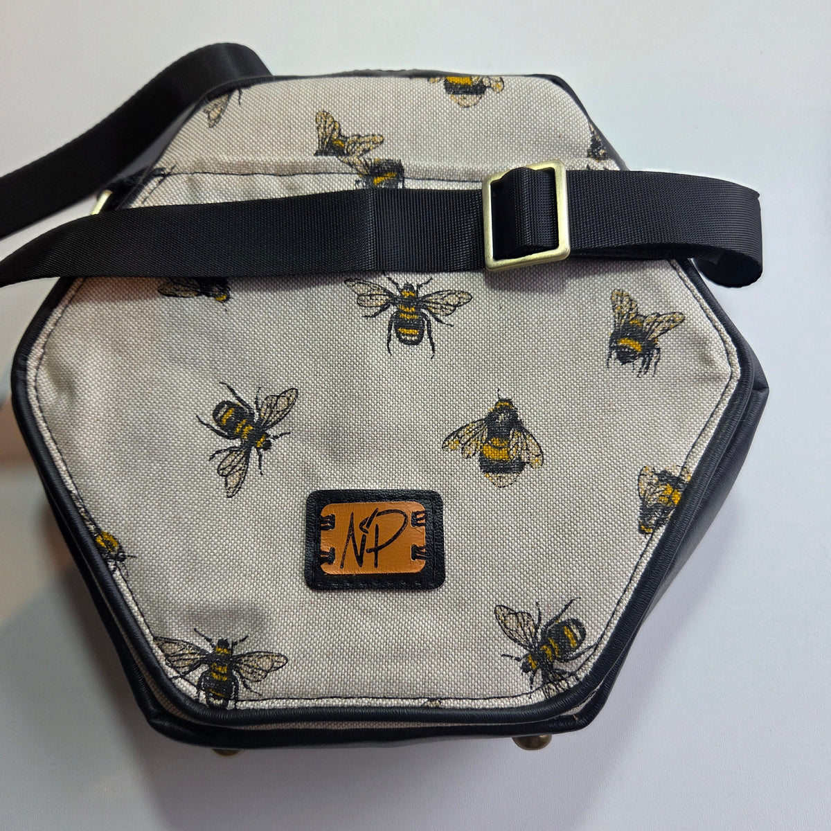Hexagonal shoulder bag with a cream background, black trim, and a black adjustable strap with a brass buckle. The canvas is printed with yellow and black bees, and features a small tan leather patch with a black monogram logo.