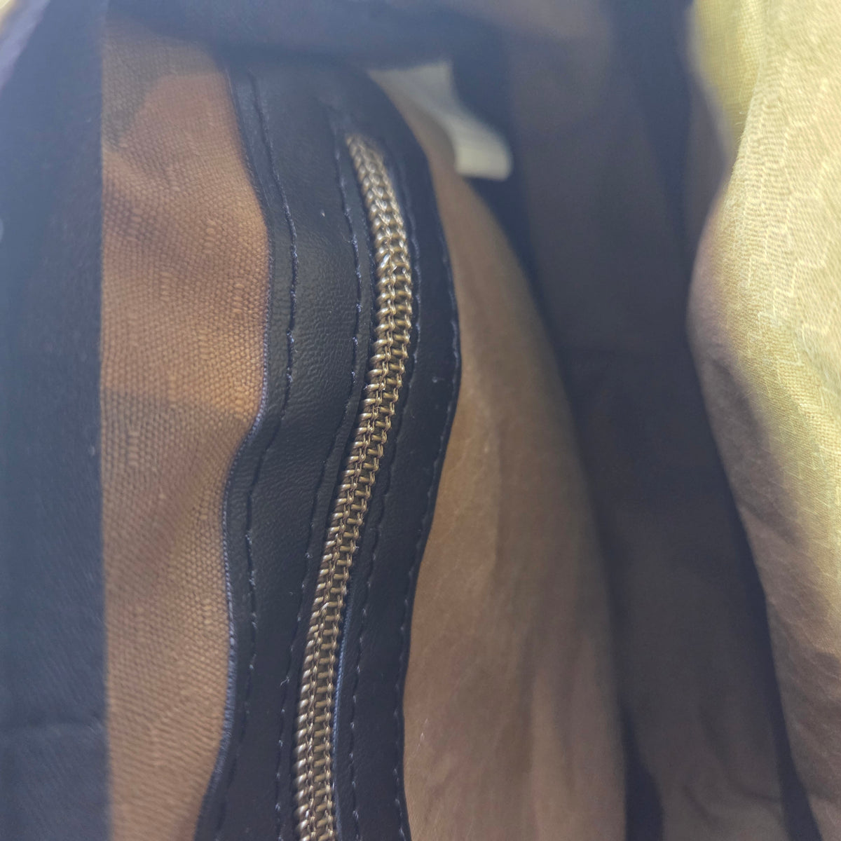 Close-up detail of a handbag's interior, showing a brass tone zipper with black vinyl trim and a dark mustard fabric lining.