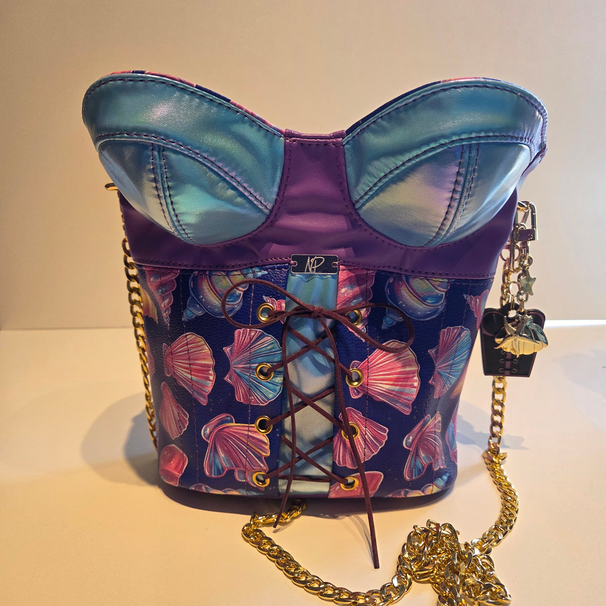A small structured handbag with a corset-inspired top, featuring iridescent blue and purple bust panels, a purple waist section, a shell print body, purple lace-up front, and gold-tone chain straps.