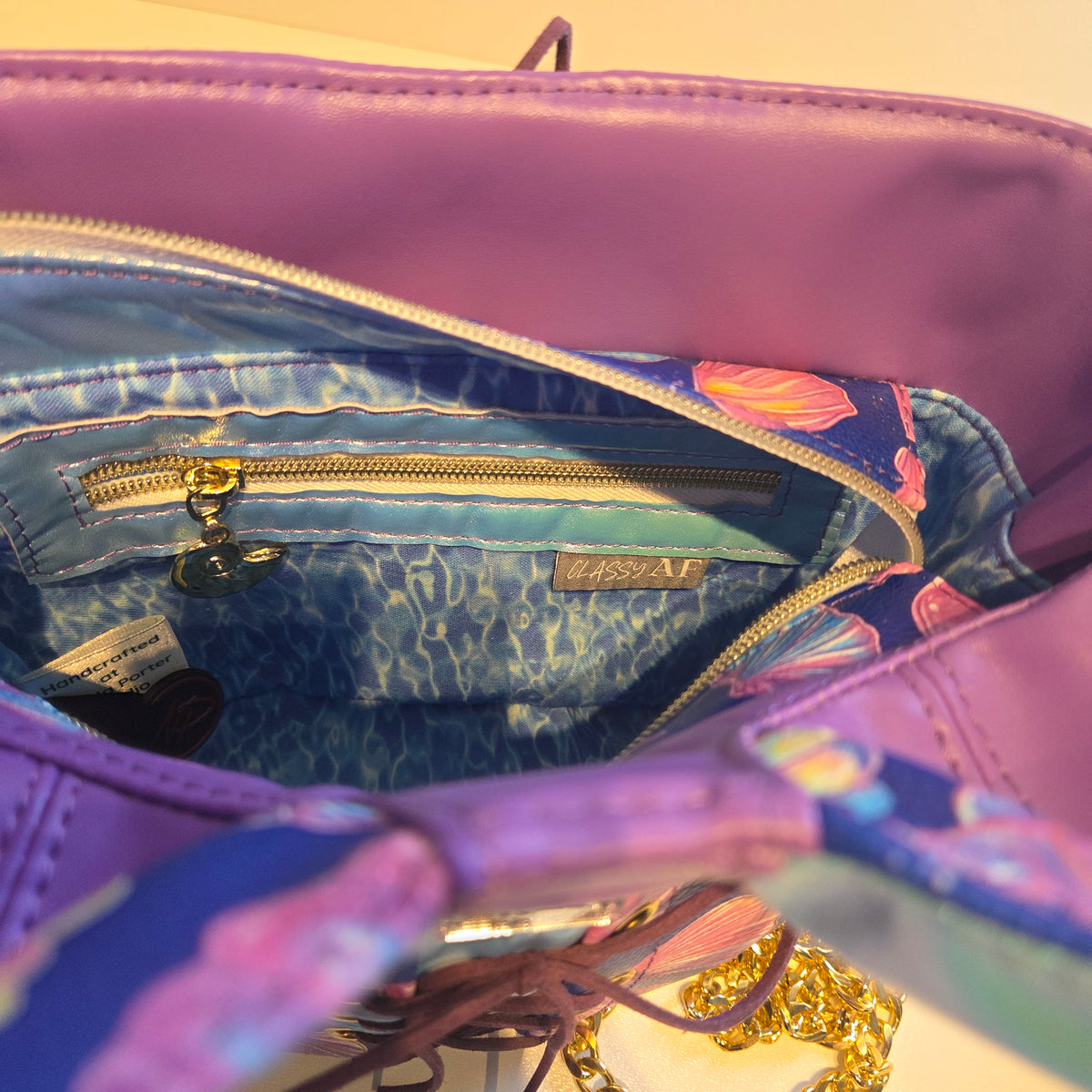 Close-up of a purple and blue patterned handbag interior showing a zippered pocket with gold sea shell hardware and a small "Classy AF" label.