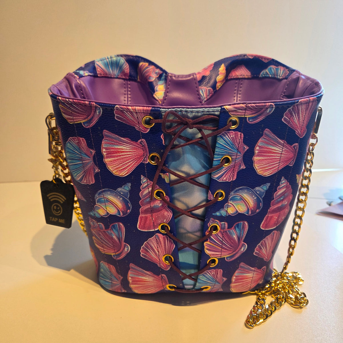 A purple and blue bag back with a pink and blue seashell pattern, featuring a front lace-up corset design with gold-tone eyelets and a gold chain strap.