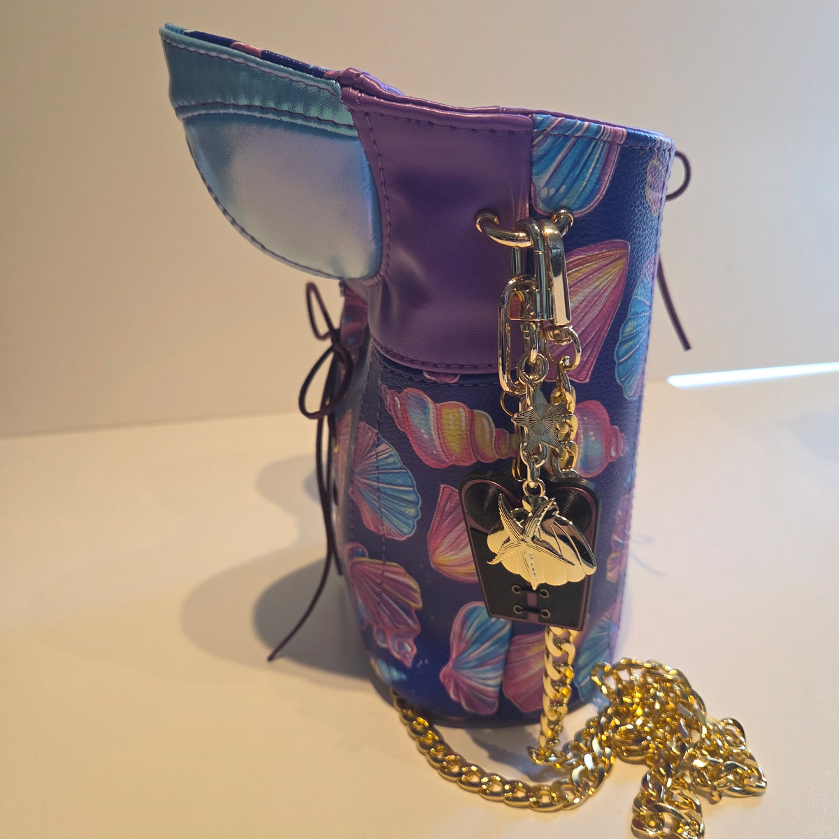 A purple and blue patterned handbag with shell motifs, featuring a gold chain strap, a black and gold charms