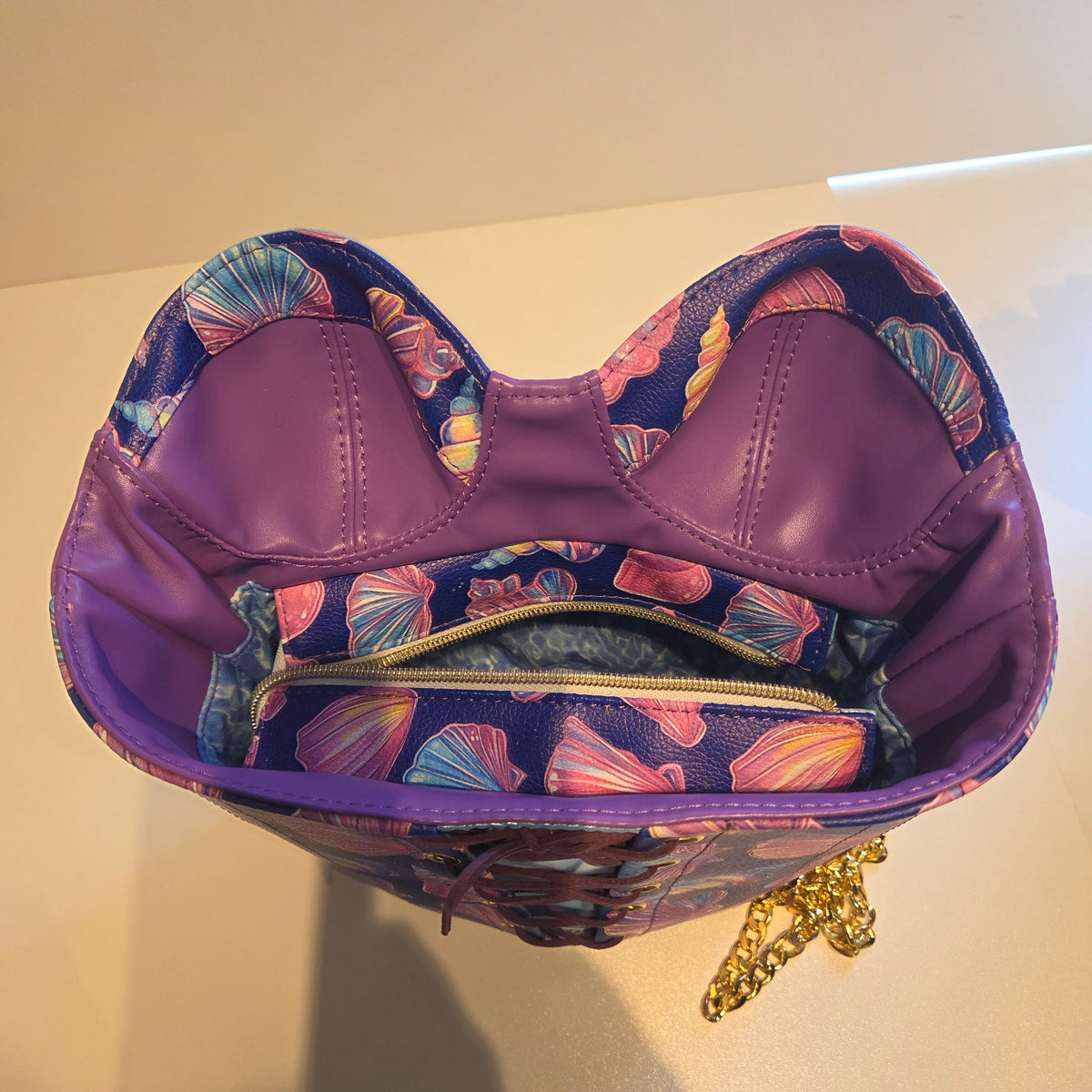 A purple bustier-style handbag with a shell-patterned exterior and a matching zippered closure, shown from a top-down angle on a light beige surface.