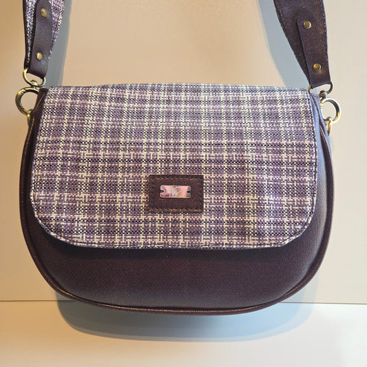 A small shoulder bag featuring a purple plaid patterned front flap and purple vinyl body and double sided strap.