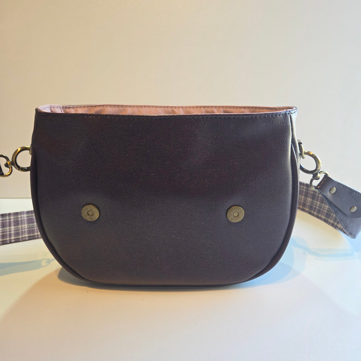A purple vinyl shoulder bag with a curved bottom, two brass snap buttons, and a plaid patterned strap, shown against a plain light background.