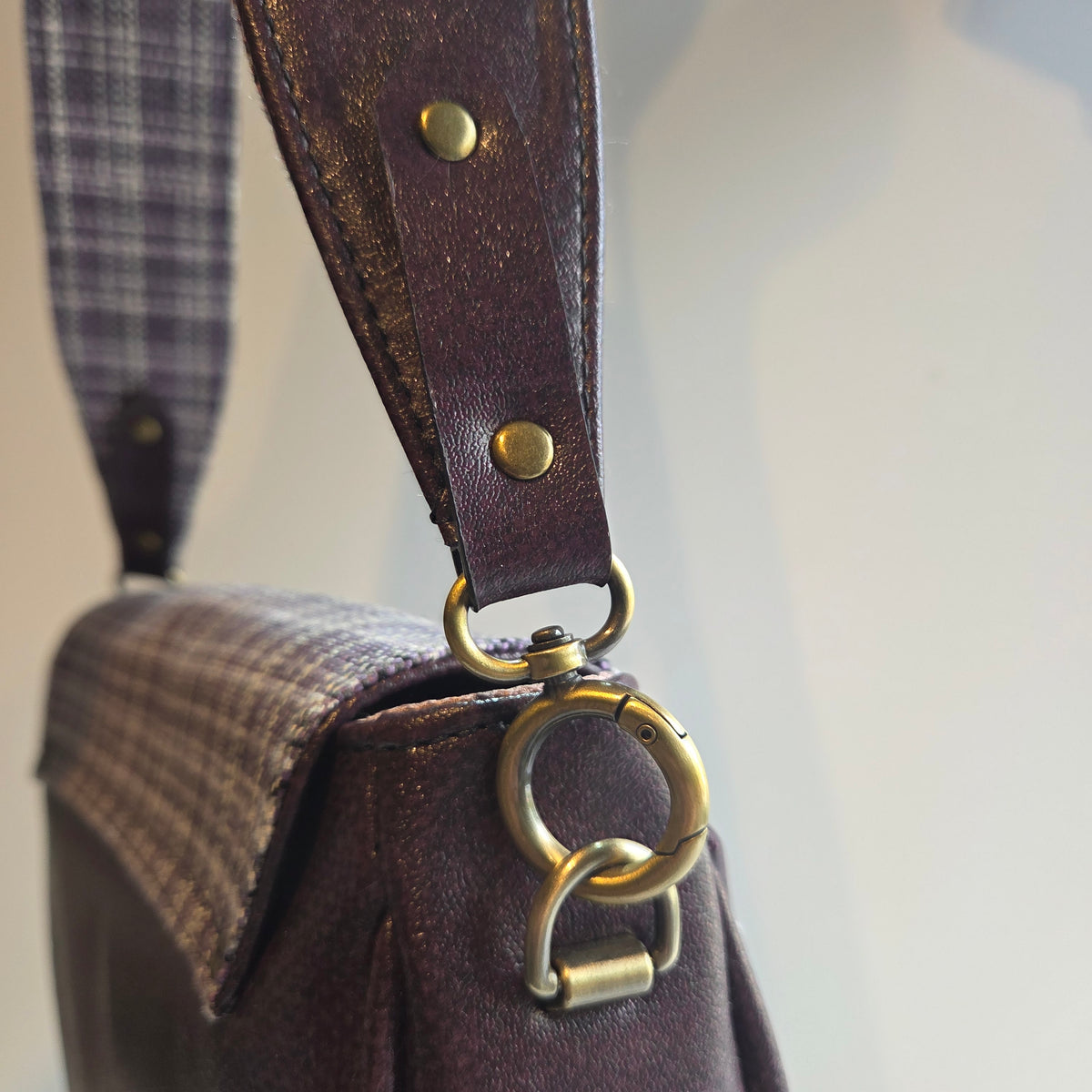 Close-up of a purple vinyl shoulder strap with antique brass hardware and a plaid fabric section, attached to a purple vinyl bag.