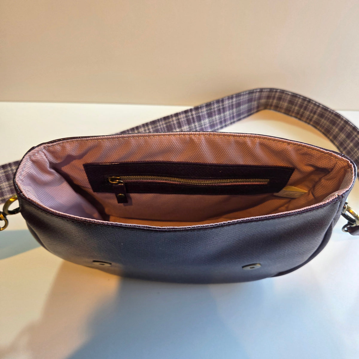 A close-up of an open purple shoulder bag showing its interior, which features a pink lining, a zippered pocket, and a plaid patterned strap.
