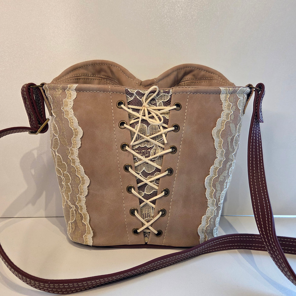 back view of a Dusky pink faux-suede bag with a lace-up corset style, cream lace panels, and a dark burgundy shoulder strap, shown against a plain off-white background.
