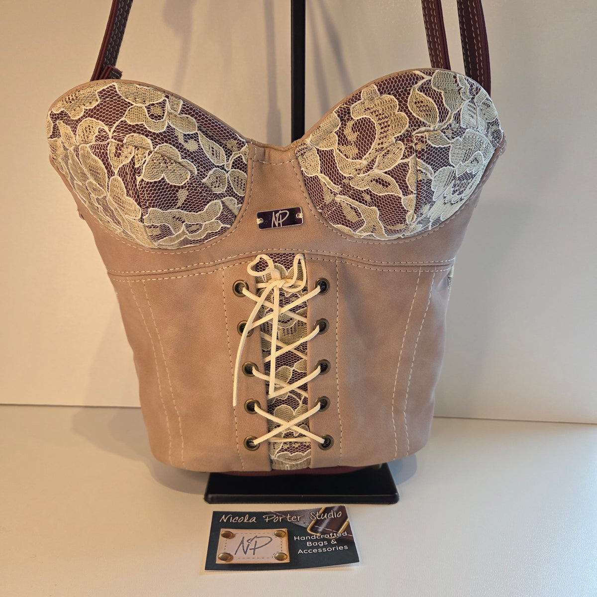 A structured corset-style handbag in dusky pink suede with cream floral lace panels on the upper sides and cream lace-up front detailing, displayed on a black stand against a plain light background.